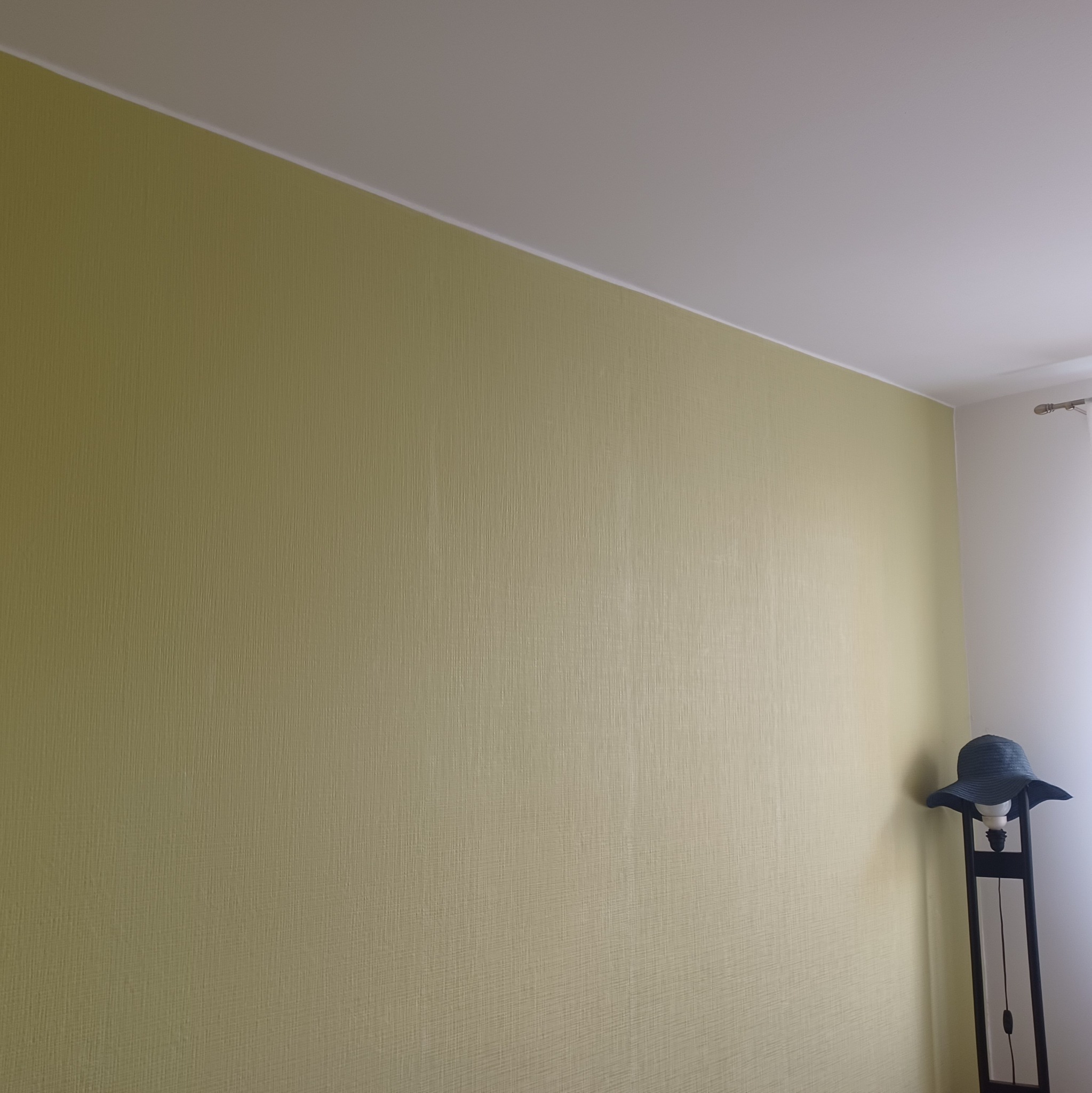 Ściana pokryta tapetą o delikatnej fakturze w odcieniach oliwkowej zieleni, widoczny fragment sufitu i narożnik pokoju z lampą podłogową, na której leży damski kapelusz.