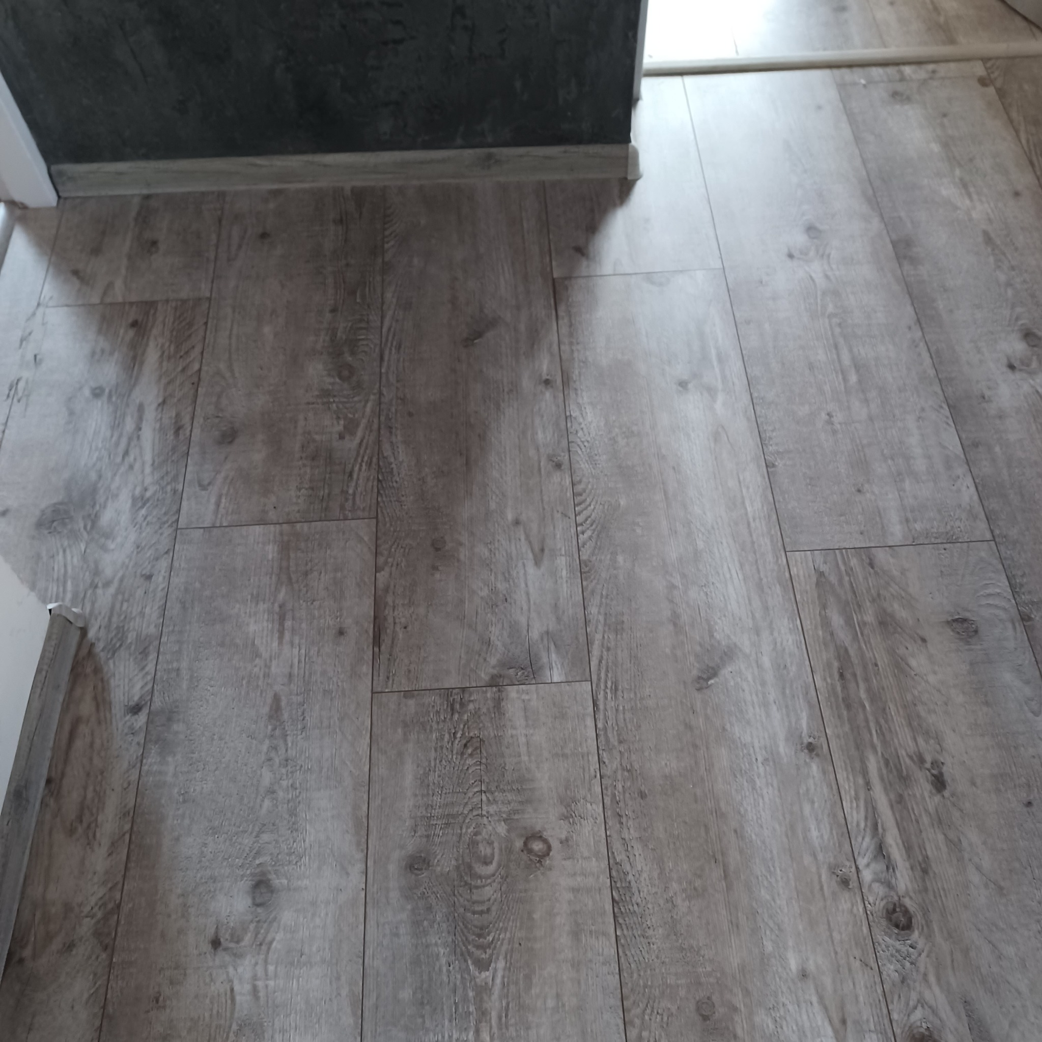 Ułożone panele podłogowe w odcieniach szarości, widoczne łączenia i faktura drewna, z fragmentem ściany w tle.