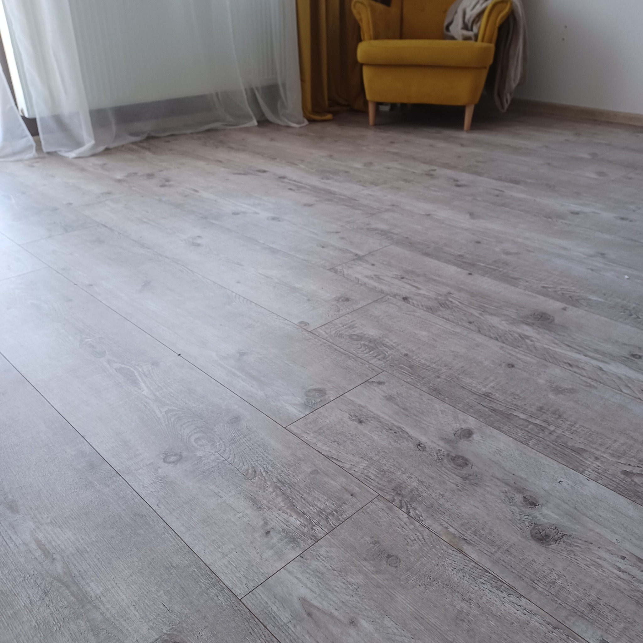 Jasnoszare panele podłogowe ułożone w jodełkę, z widocznym żółtym fotelem i zasłonami w tle.