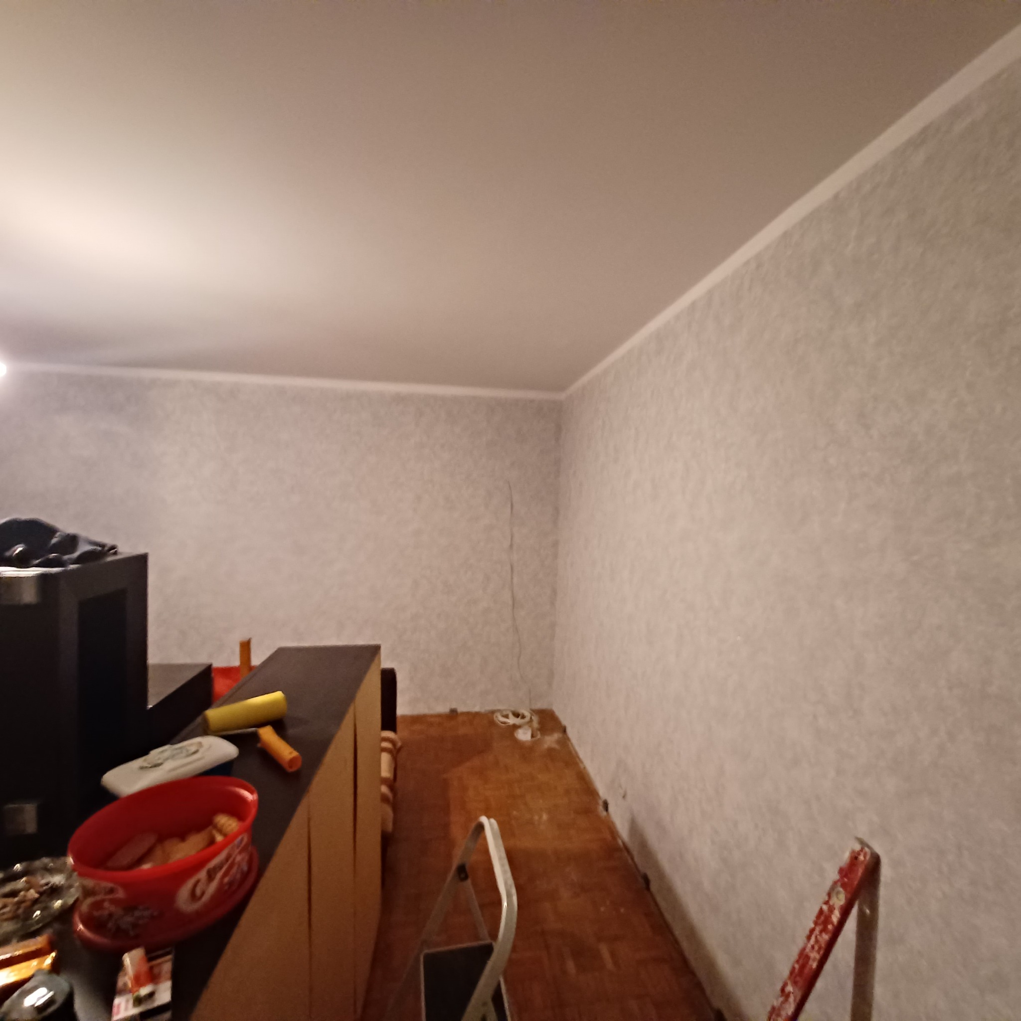 Pokój w trakcie remontu: ściany pokryte tapetą o delikatnym wzorze, na podłodze częściowo widoczne stare panele, drabina malarska, wałki i kuweta z farbą na meblach.