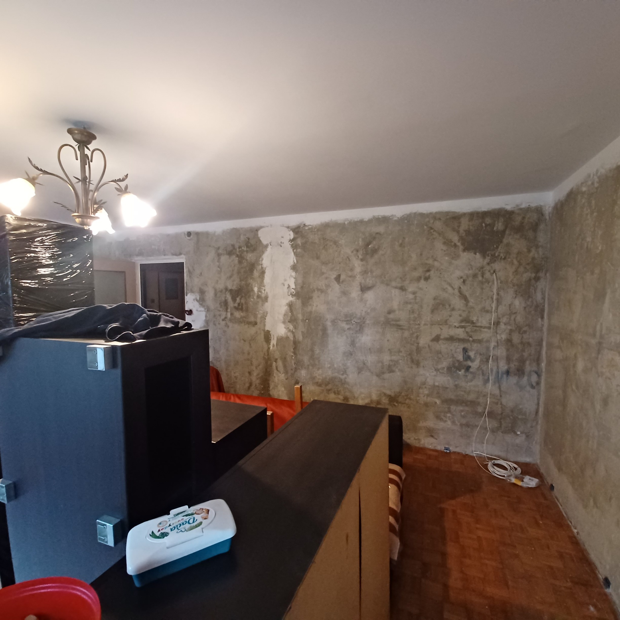 Pokój w trakcie remontu: ściany przygotowane do malowania lub tapetowania, widoczne ślady po szpachlowaniu, podłoga częściowo zakryta meblami, wisząca lampa sufitowa.