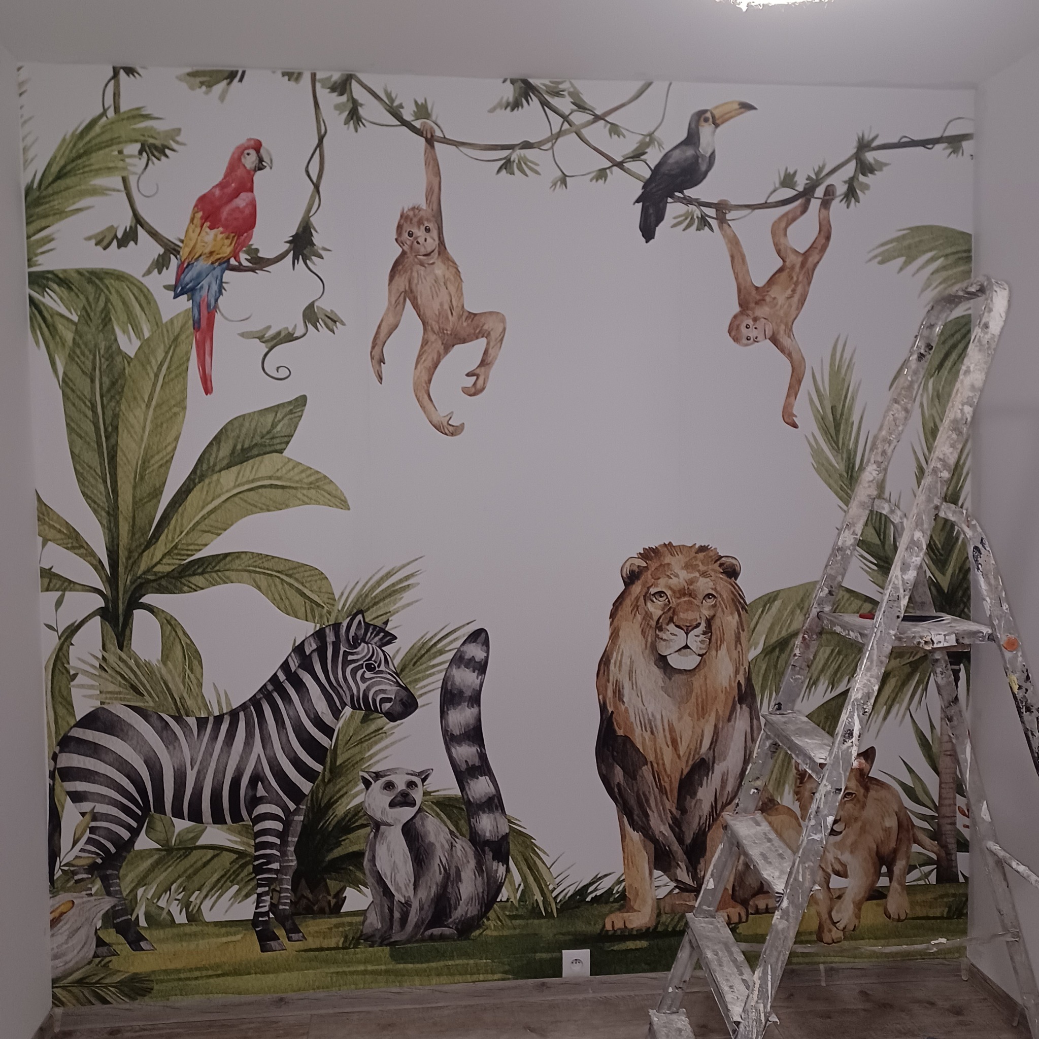 Tapeta z motywem dżungli z akwarelowymi zwierzętami, takimi jak lew, zebra, lemur, małpy, tukan i papuga, częściowo zasłonięta przez aluminiową drabinę malarską.