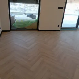 Ułożone panele podłogowe w jodełkę francuską, widok z góry, jasne drewno. Nowoczesne wnętrze w Poniecu z dużymi oknami i widokiem na zewnątrz.