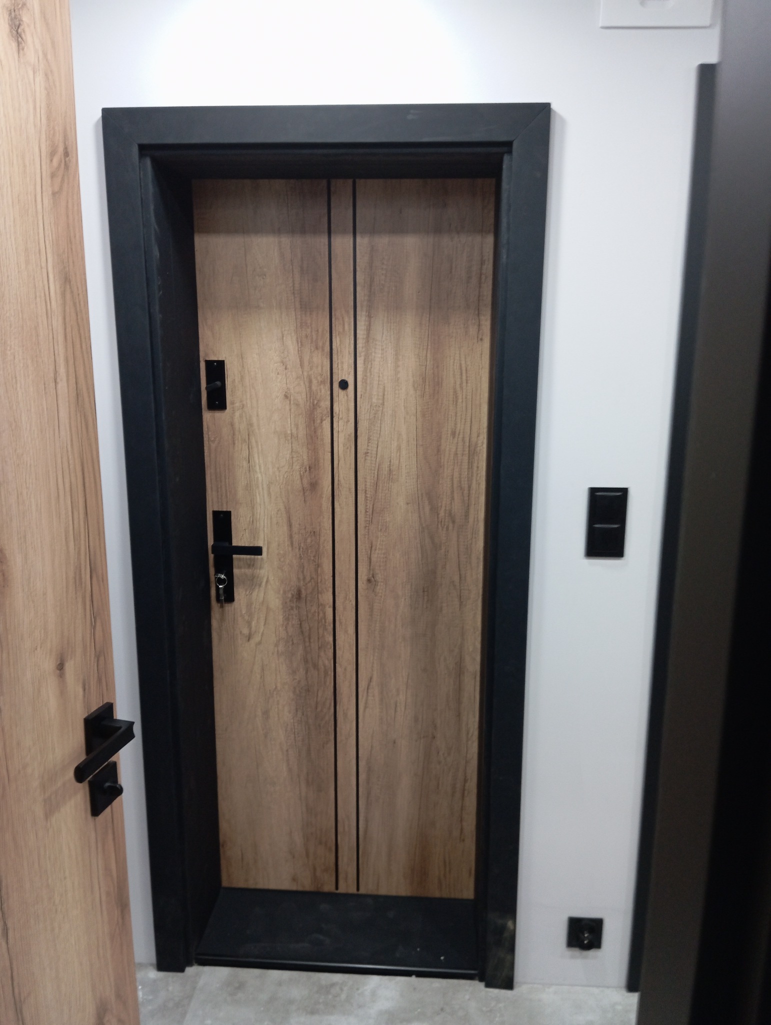 Drewniane drzwi wewnętrzne z czarną ramą i okuciami, widoczne w bliskim ujęciu z fragmentem sąsiednich drzwi o podobnym wykończeniu.