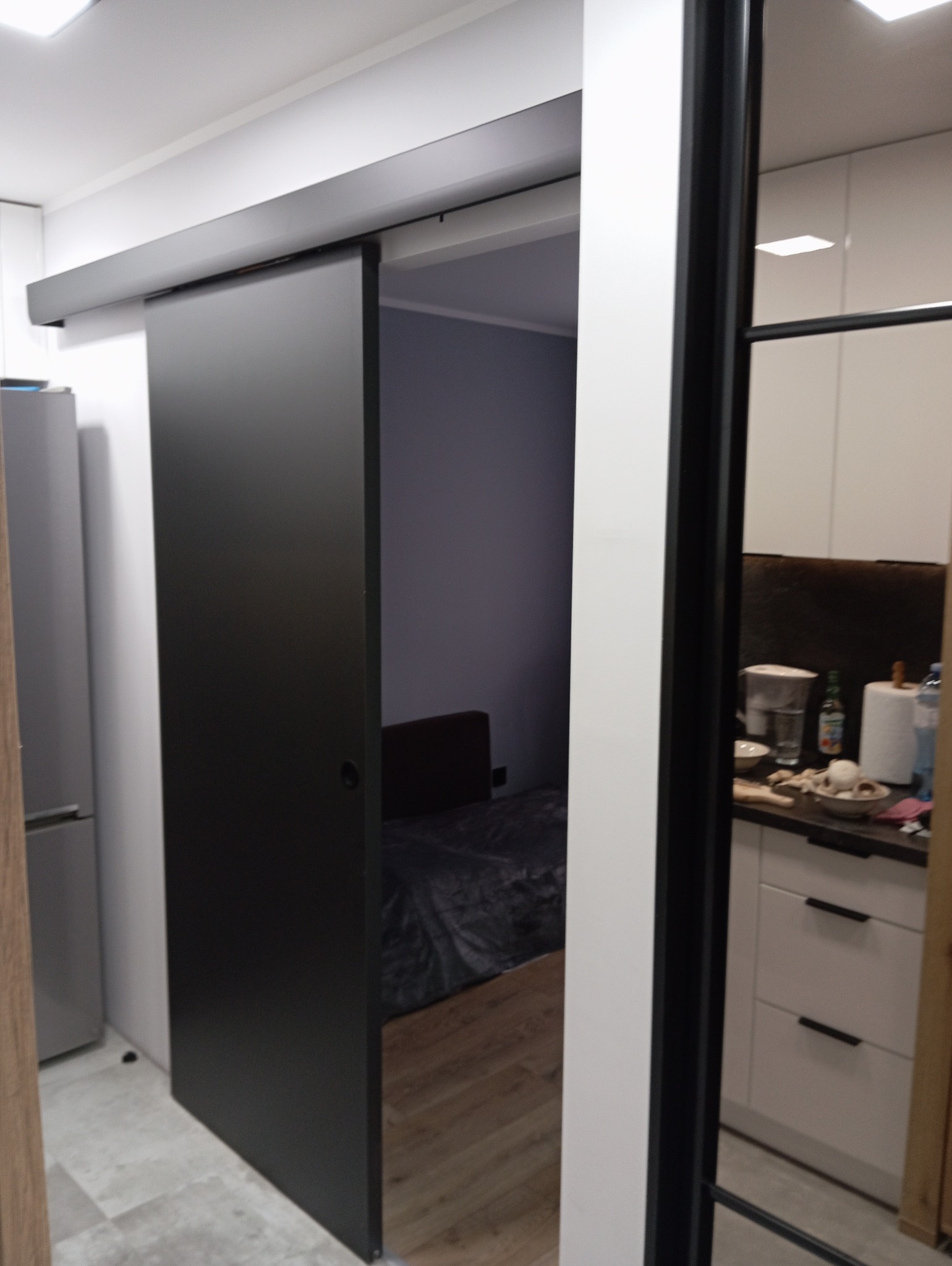 Czarne przesuwne drzwi wewnętrzne w nowoczesnym mieszkaniu, widok na sypialnię z parkietem i kuchnię z białymi szafkami.