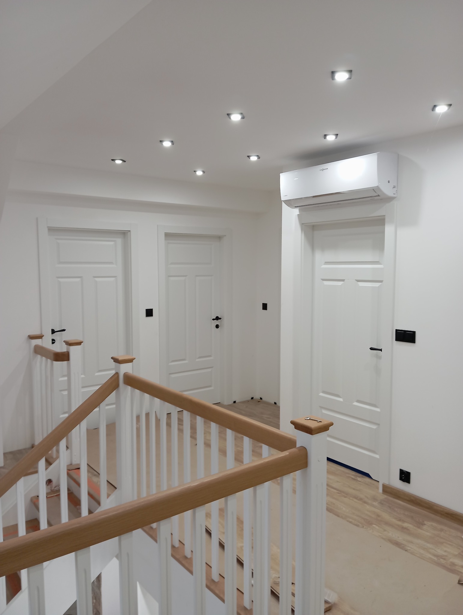 Wnętrze domu w trakcie wykańczania: widok na korytarz z trzema białymi drzwiami z czarnymi klamkami, białą balustradę schodów z drewnianymi elementami oraz zamontowany klimatyzator na ścianie...