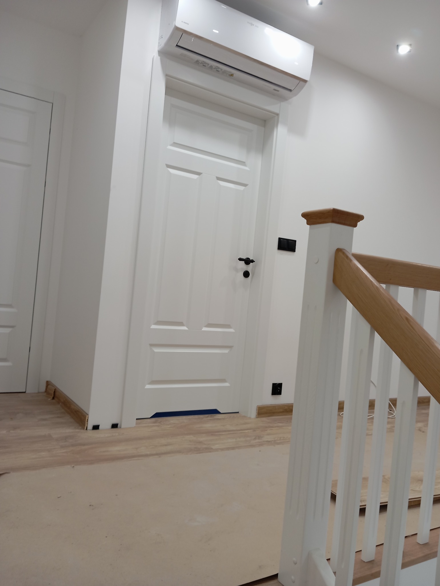 Białe, panelowe drzwi wewnętrzne z czarną klamką, zamontowane w białej ścianie, nad drzwiami klimatyzator, widoczny fragment drewnianej balustrady schodów, podłoga częściowo zabezpieczona kartonem.