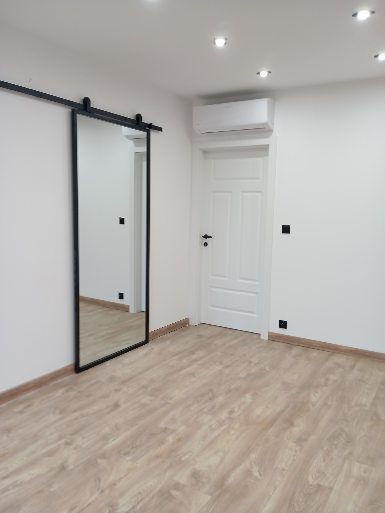 Wnętrze pokoju z przesuwnymi drzwiami lustrzanymi na czarnej szynie, białą ścianą, drzwiami i panelami podłogowymi w odcieniach beżu.