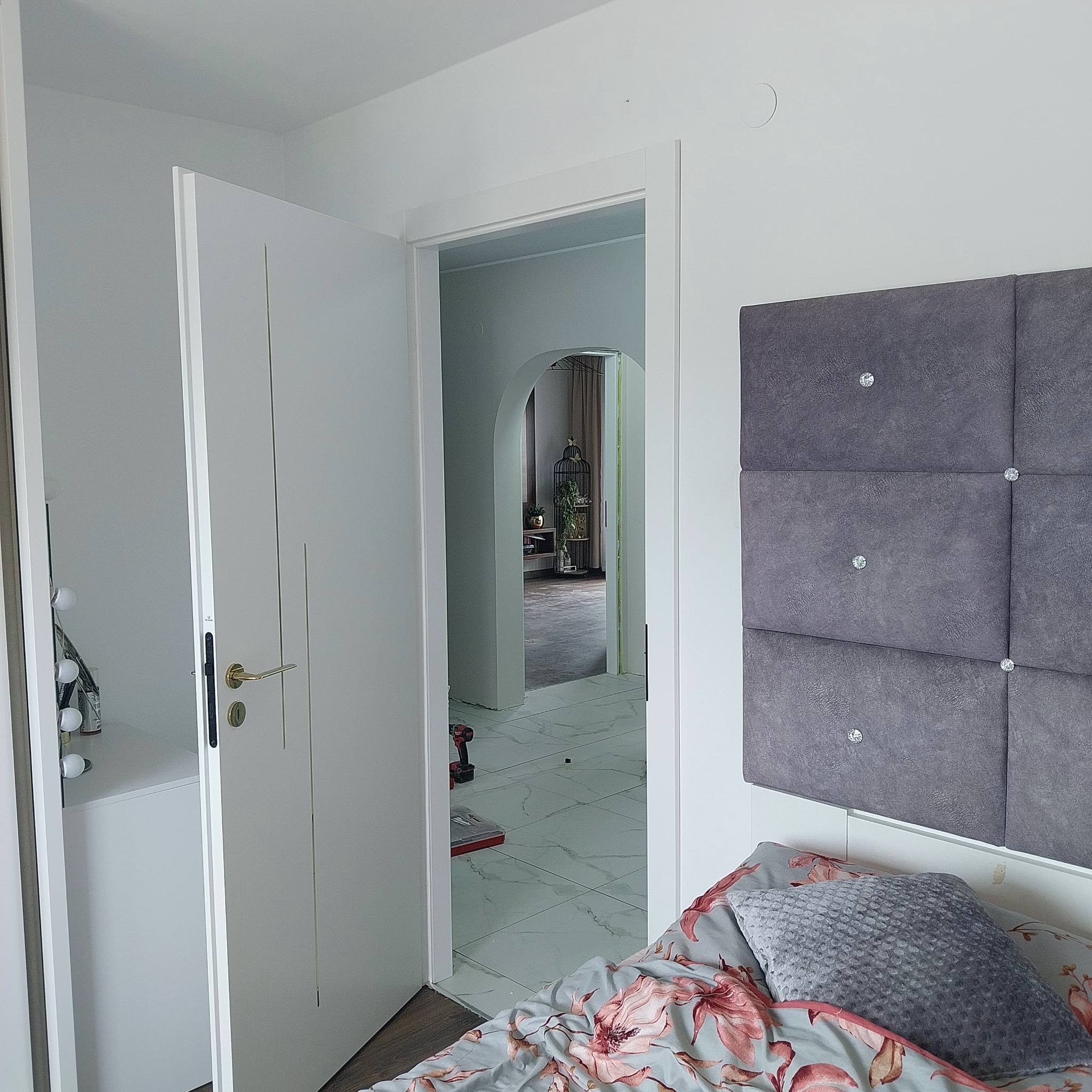 Białe drzwi wewnętrzne z złotą klamką, widoczne przez otwarte drzwi narzędzia i biała podłoga z wzorem marmuru, w tle dekoracyjna klatka z roślinami.