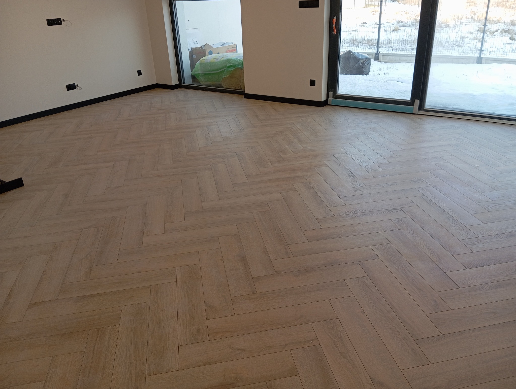 Jasne panele ułożone w jodełkę, widok na duże okno z zasypanym śniegiem ogrodem. Minimalistyczne wnętrze, czarne listwy przypodłogowe.