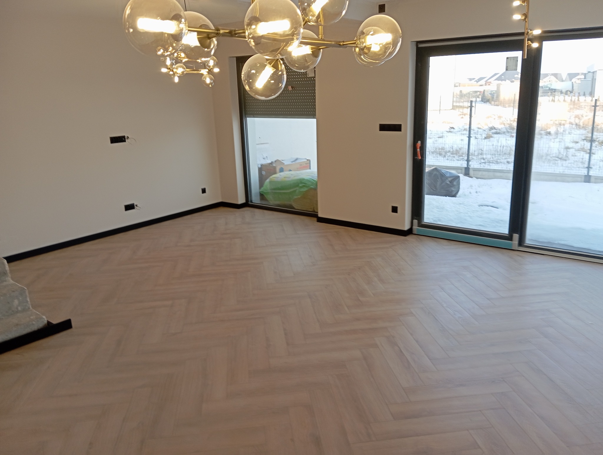 Jasne panele ułożone w jodełkę francuską w przestronnym, słonecznym salonie z dużymi oknami. Widoczne czarne listwy przypodłogowe i nowoczesna lampa sufitowa.