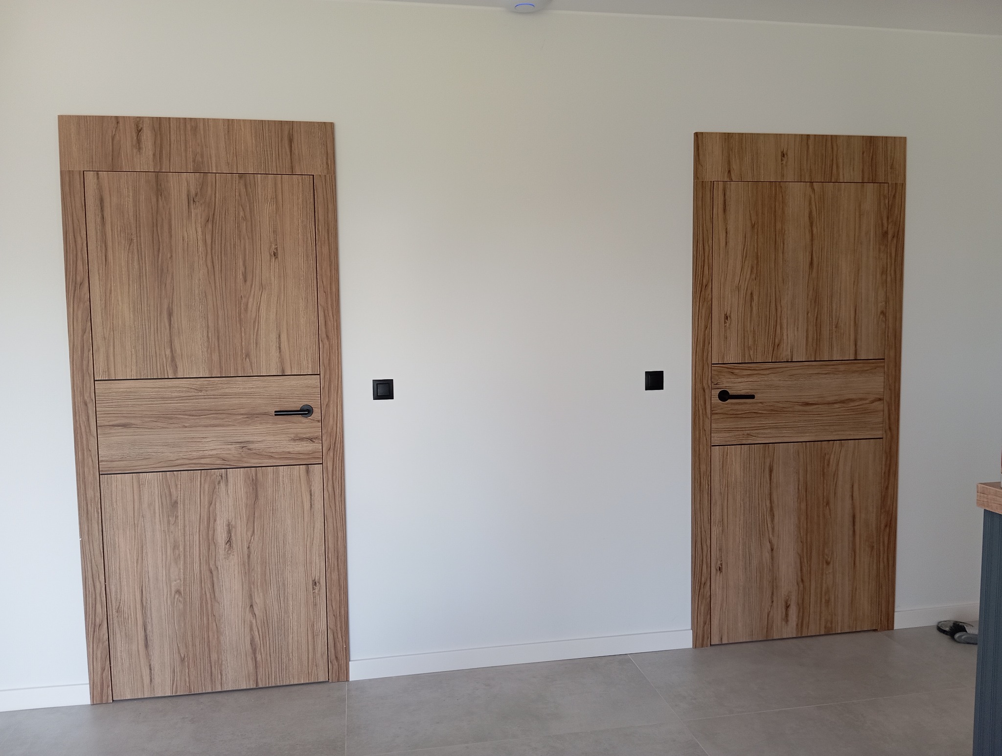 Dwa drewniane drzwi wewnętrzne z czarnymi klamkami zamontowane w białej ścianie. Widoczny fragment podłogi i listwy przypodłogowe.