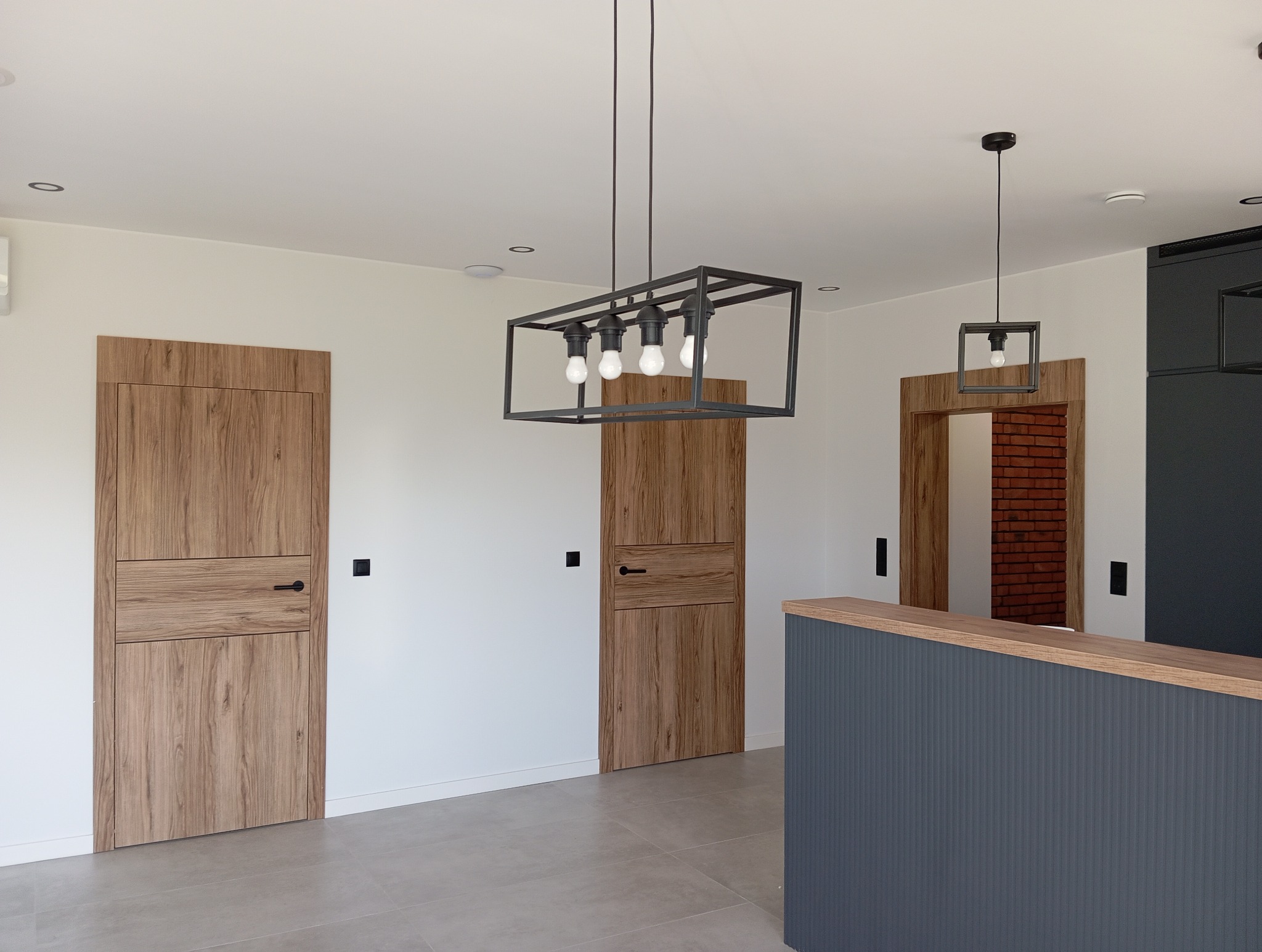 Wnętrze z dwiema drewnianymi drzwiami z poziomymi panelami, nowoczesnym oświetleniem i szarą zabudową. Widok na ceglaną ścianę przez otwarte drzwi.