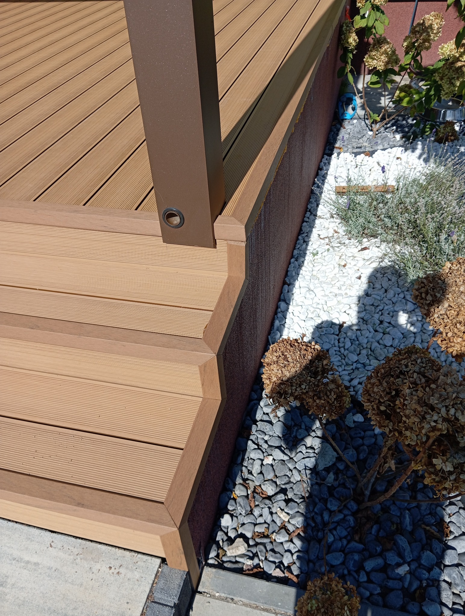 Fragment tarasu kompozytowego z brązowym słupkiem, widoczne deski i obrzeża. Obok rabata z białymi i szarymi kamieniami oraz roślinnością.