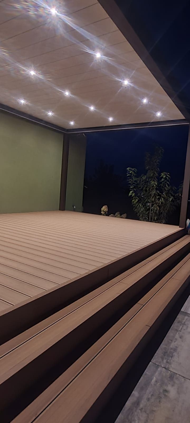 Oświetlony taras kompozytowy z drewnianymi schodami w nocy, widok z dołu, zieleń w tle.