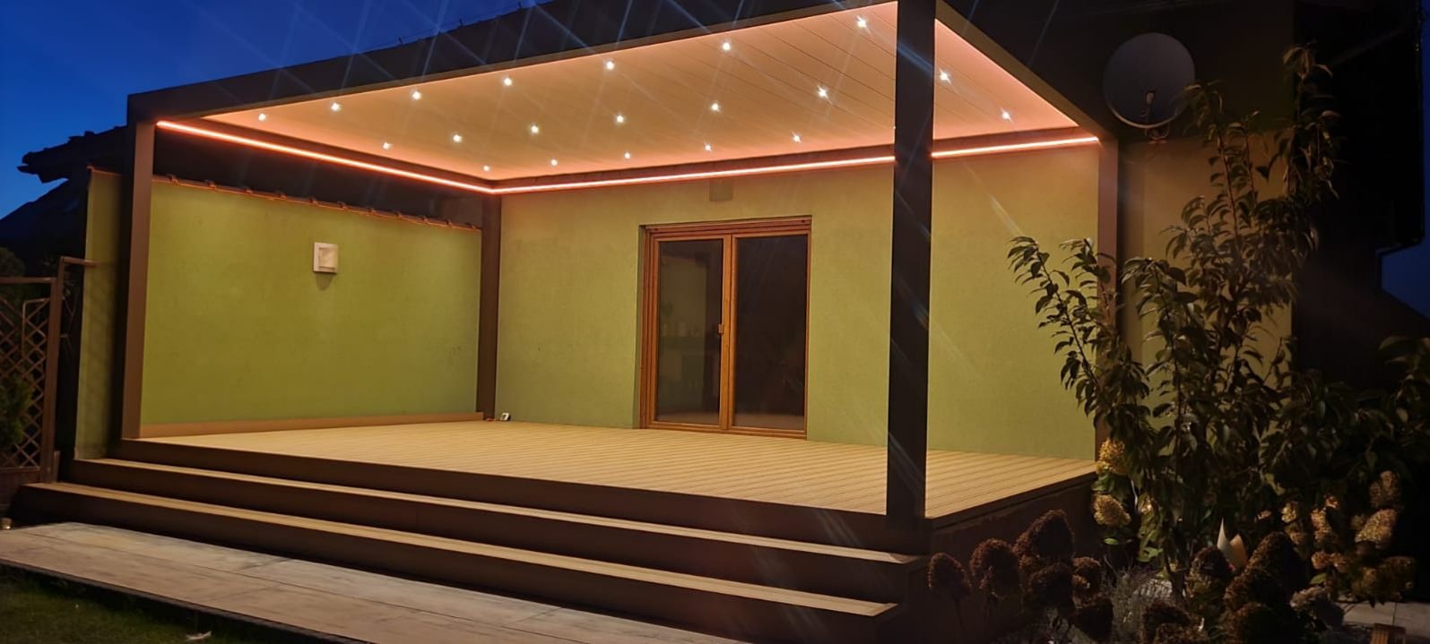 Nowoczesny taras kompozytowy z oświetleniem LED, widok nocny. Drewnopodobna podłoga, minimalistyczna konstrukcja z zadaszeniem i roślinność w tle.