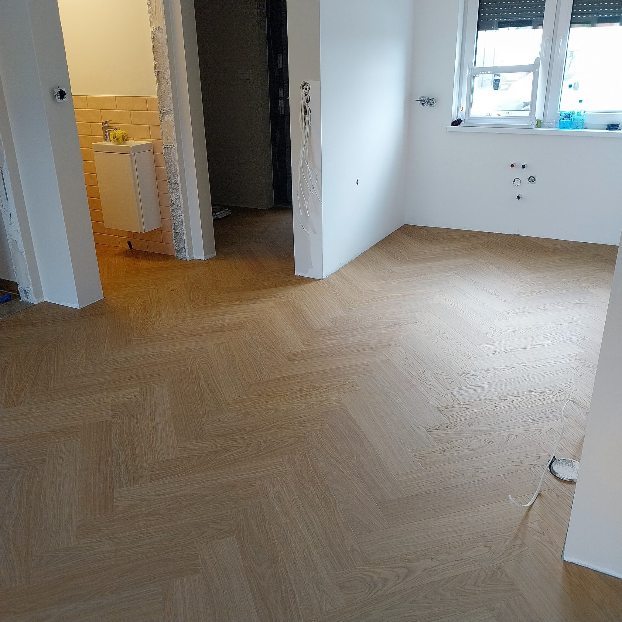 Ukończona podłoga z paneli ułożonych w jodełkę, widok na łazienkę i pokój z oknem. Jasne panele i białe ściany tworzą nowoczesny wygląd wnętrza.