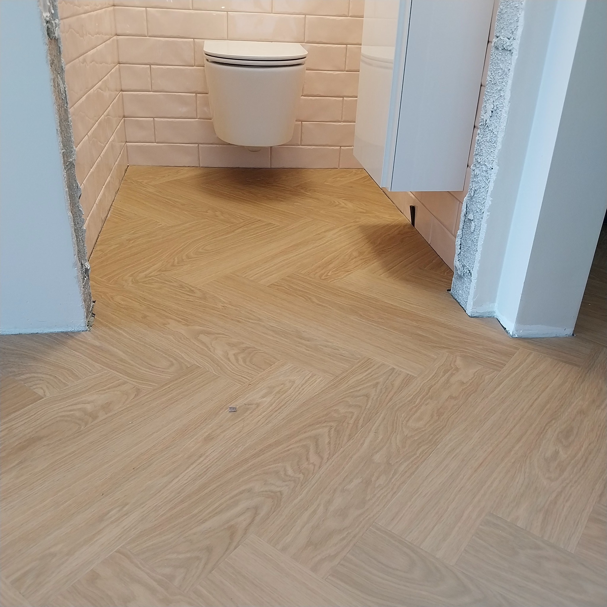 Jasne panele podłogowe ułożone we wzór jodełki w wykończonej łazience z podwieszaną toaletą i białą szafką. Widoczne niedokończone wykończenia ścian.