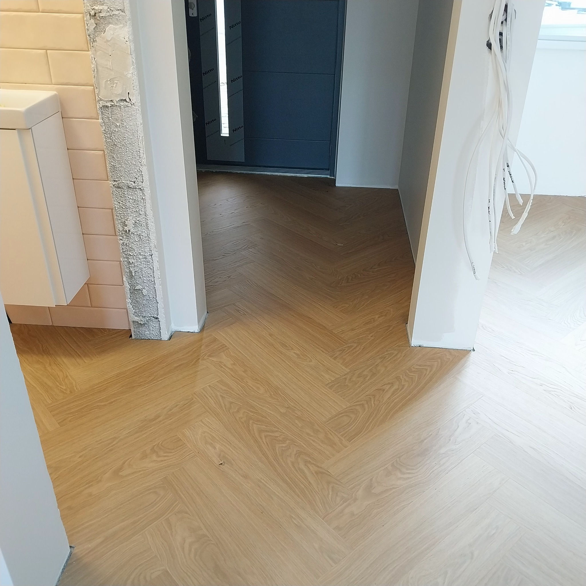 Jasne panele ułożone w jodełkę w korytarzu, widok na drzwi wejściowe i niedokończone wykończenie ścian, z widocznymi przewodami elektrycznymi.