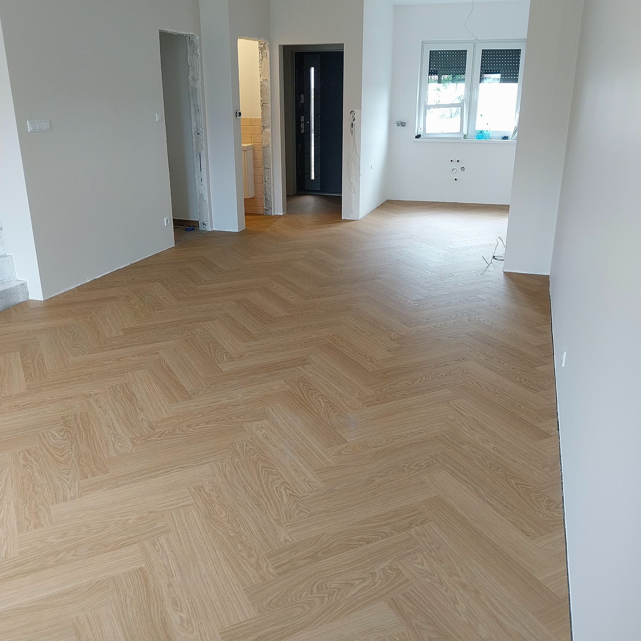 Nowoczesne wnętrze z ułożonymi panelami w jodełkę. Widoczne drzwi wejściowe i okno z roletami. Świeżo wykończone ściany. Minimalistyczny design.