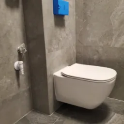Nowoczesna podwieszana toaleta w łazience wykończonej szarymi płytkami imitującymi kamień, widoczny niebieski element instalacji oraz zaw&oacute;r dopływowy wody.
