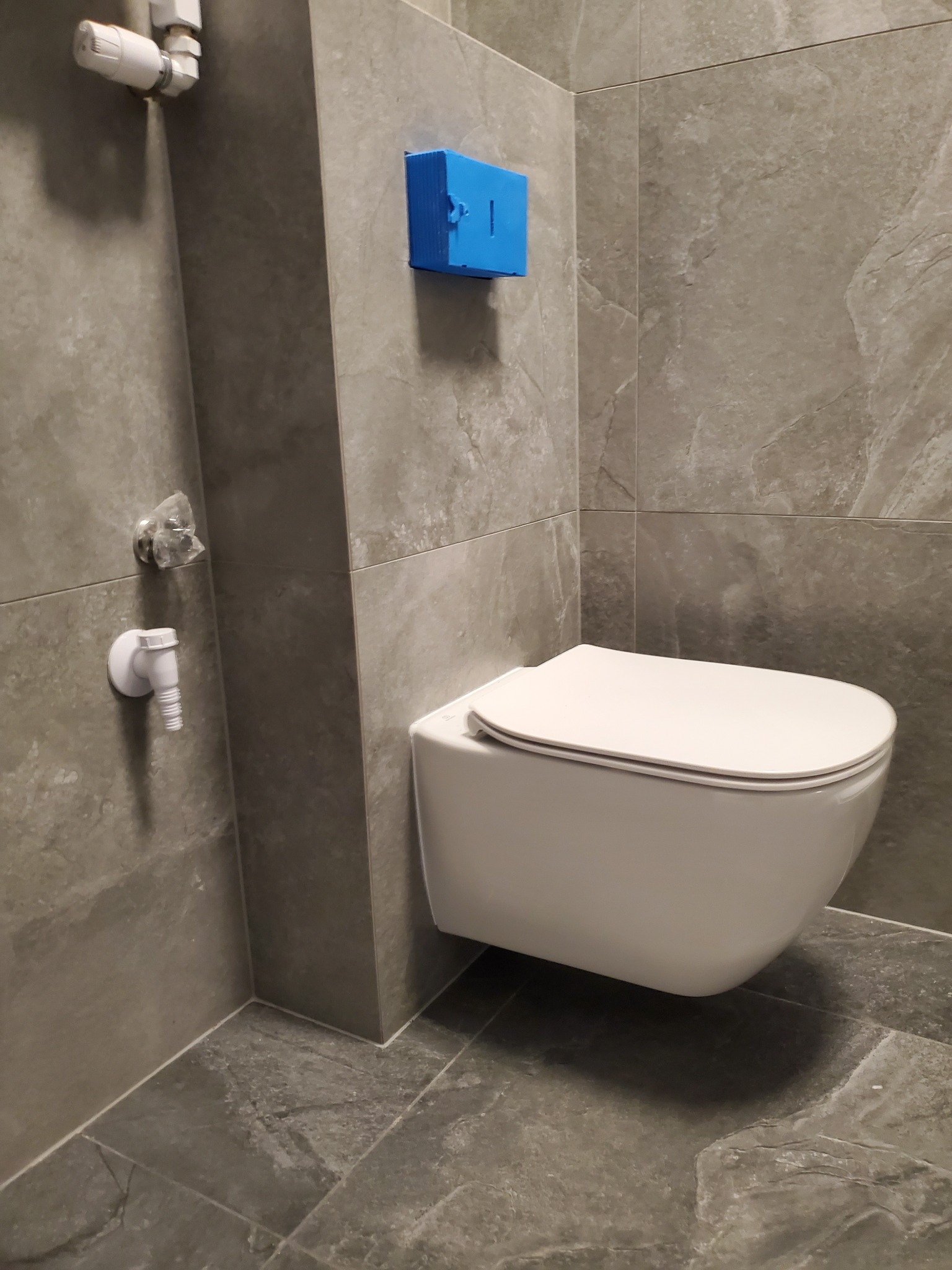 Nowoczesna podwieszana toaleta w łazience wykończonej szarymi płytkami imitującymi kamień, widoczny niebieski element instalacji oraz zawór dopływowy wody.