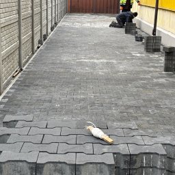 KANBRUK Patryk Kudrzycki - Prace brukarskie w toku: dwóch robotników układa szarą kostkę brukową o nietypowym kształcie na długim podjeździe, stosy kostki czekają na ułożenie, widoczna sznurkowa linia wyznaczająca kierunek...