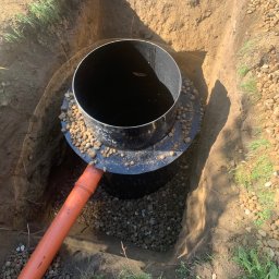 KANBRUK Patryk Kudrzycki - Montaż czarnego, cylindrycznego zbiornika w wykopie, obsypanego drobnymi kamieniami, z podłączoną pomarańczową rurą kanalizacyjną.