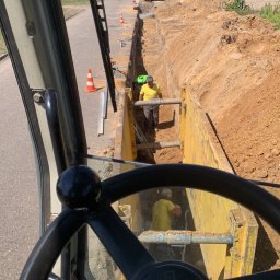 KANBRUK Patryk Kudrzycki - Widok z kabiny operatora koparki na wykop zabezpieczony żółtymi szalunkami, w którym pracują dwaj robotnicy w żółtych koszulkach i kaskach, w tle ulica i zaparkowany bus.