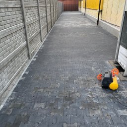 KANBRUK Patryk Kudrzycki - Ułożona szara kostka brukowa typu 'H' na posesji, widoczny fragment betonowego ogrodzenia i żółty kompresor z pomarańczowym przewodem.