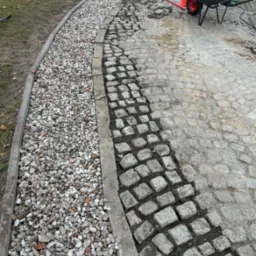 Układanie kostki brukowej: fragment chodnika z szarej kostki, częściowo ułożonej, z widoczną warstwą podsypki, obok obsypka z drobnych kamieni, w tle taczka i robotnicy.