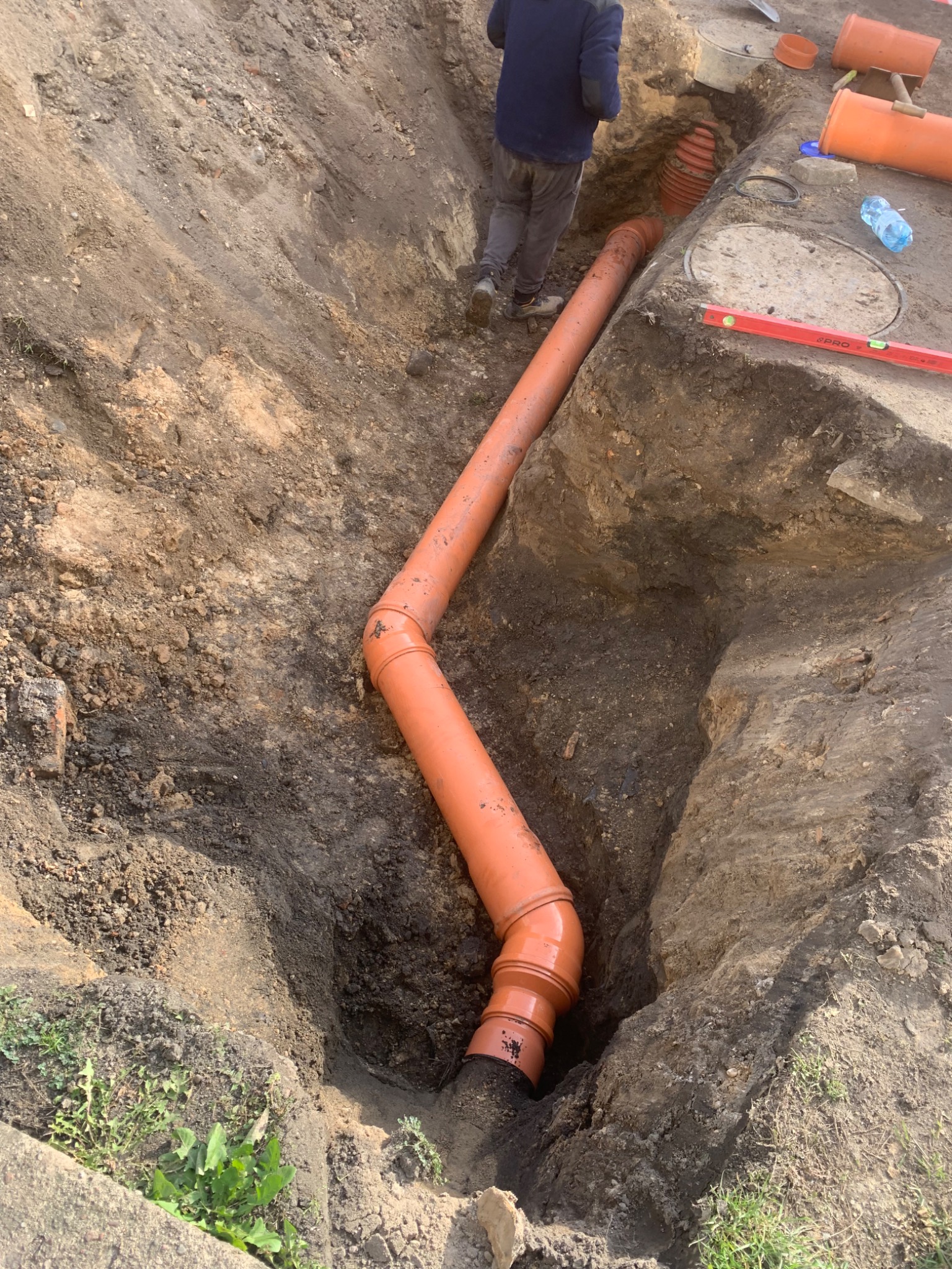 Pomarańczowa rura kanalizacyjna ułożona w wykopie ziemnym, widoczny fragment studzienki i poziomica PRO.