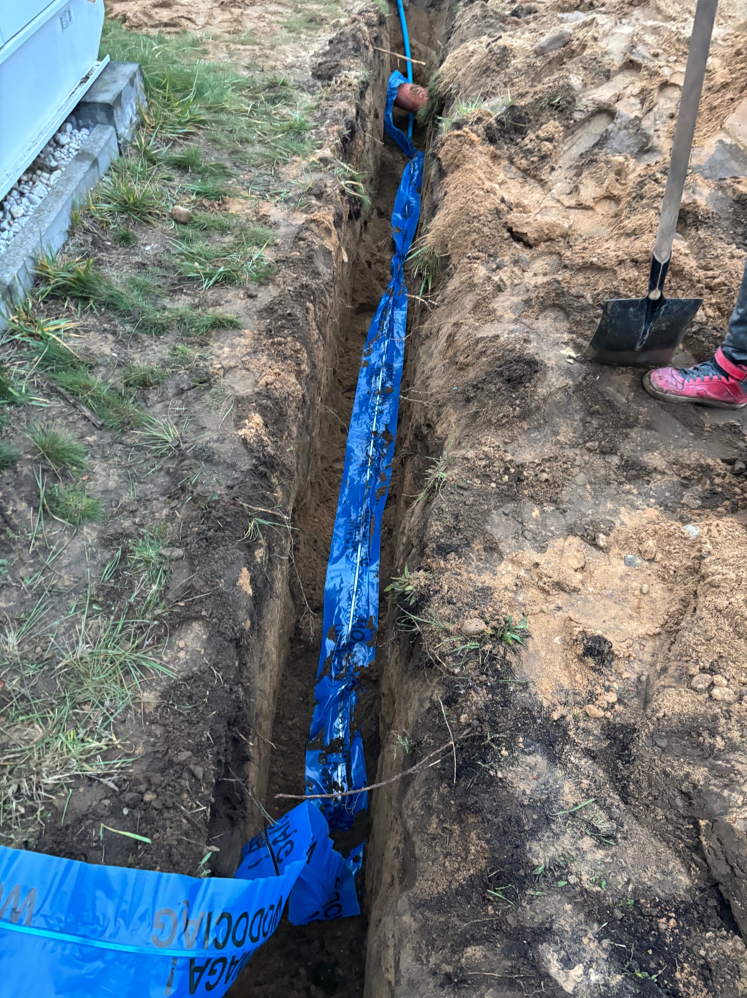 Wykop w ziemi z niebieską taśmą ostrzegawczą i rurą wodną, widoczna łopata i fragment buta osoby pracującej.