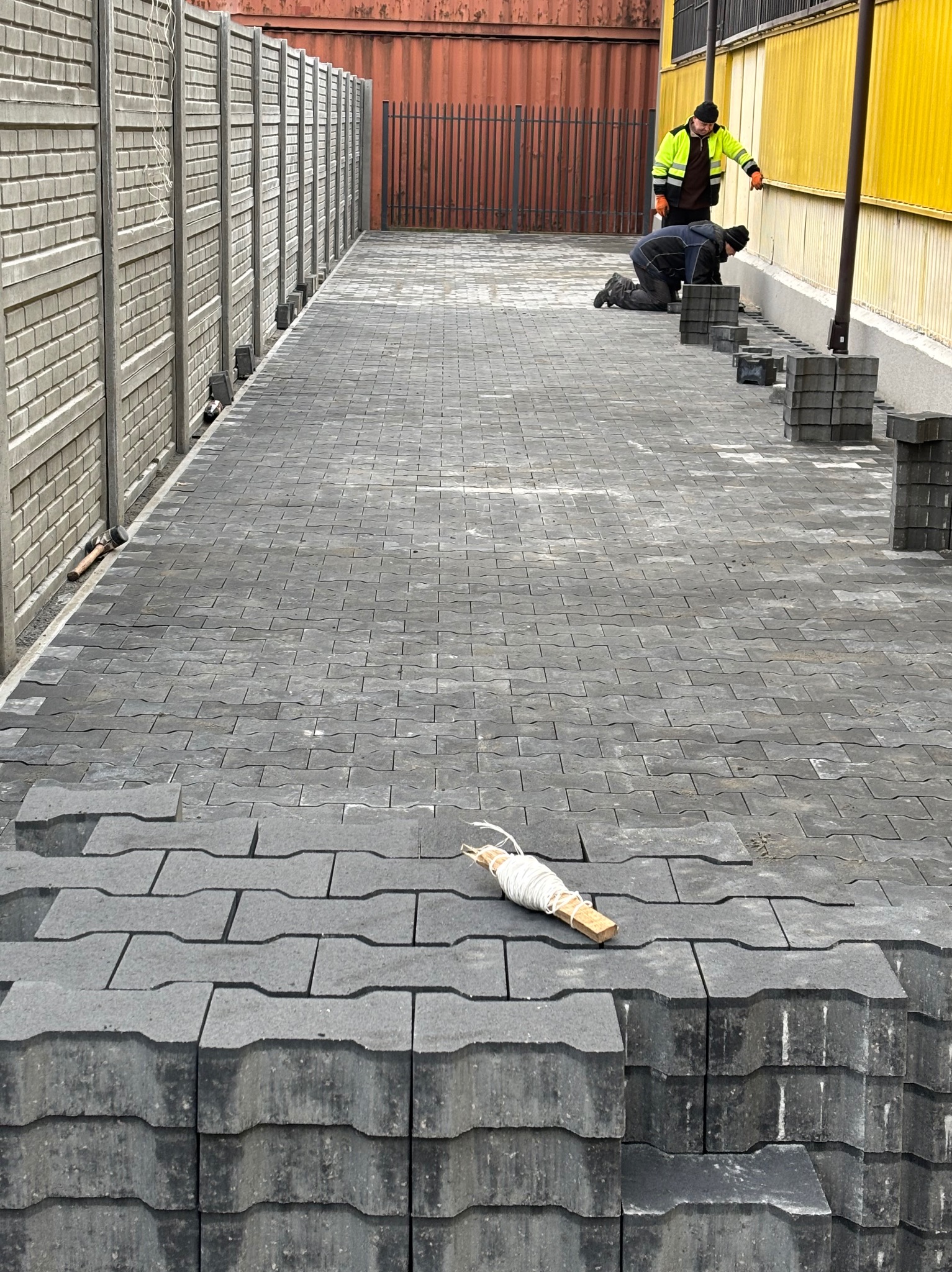 Prace brukarskie w toku: dwóch robotników układa szarą kostkę brukową o nietypowym kształcie na długim podjeździe, stosy kostki czekają na ułożenie, widoczna sznurkowa linia wyznaczająca kierunek...