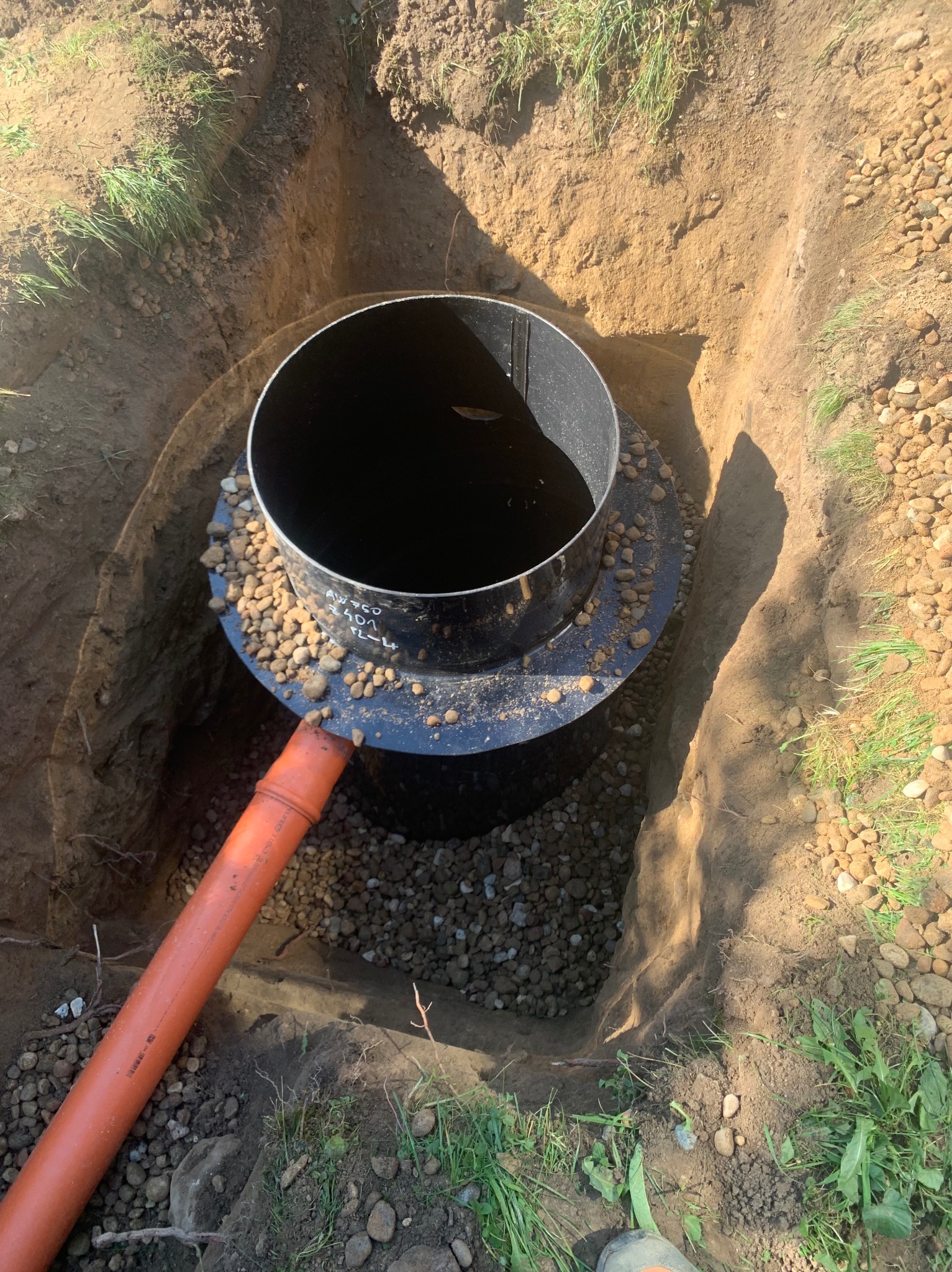 Montaż czarnego, cylindrycznego zbiornika w wykopie, obsypanego drobnymi kamieniami, z podłączoną pomarańczową rurą kanalizacyjną.
