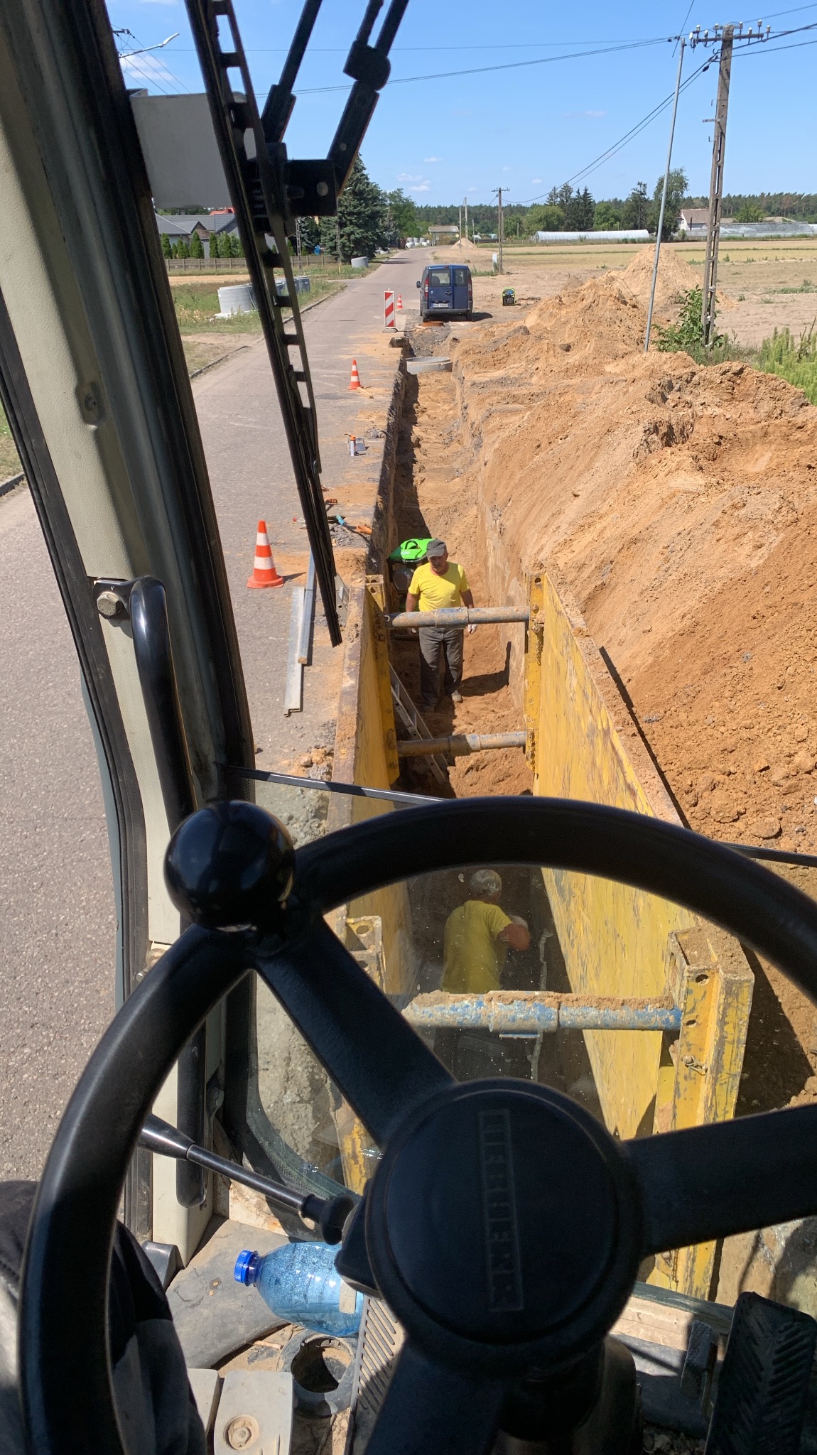 Widok z kabiny operatora koparki na wykop zabezpieczony żółtymi szalunkami, w którym pracują dwaj robotnicy w żółtych koszulkach i kaskach, w tle ulica i zaparkowany bus.