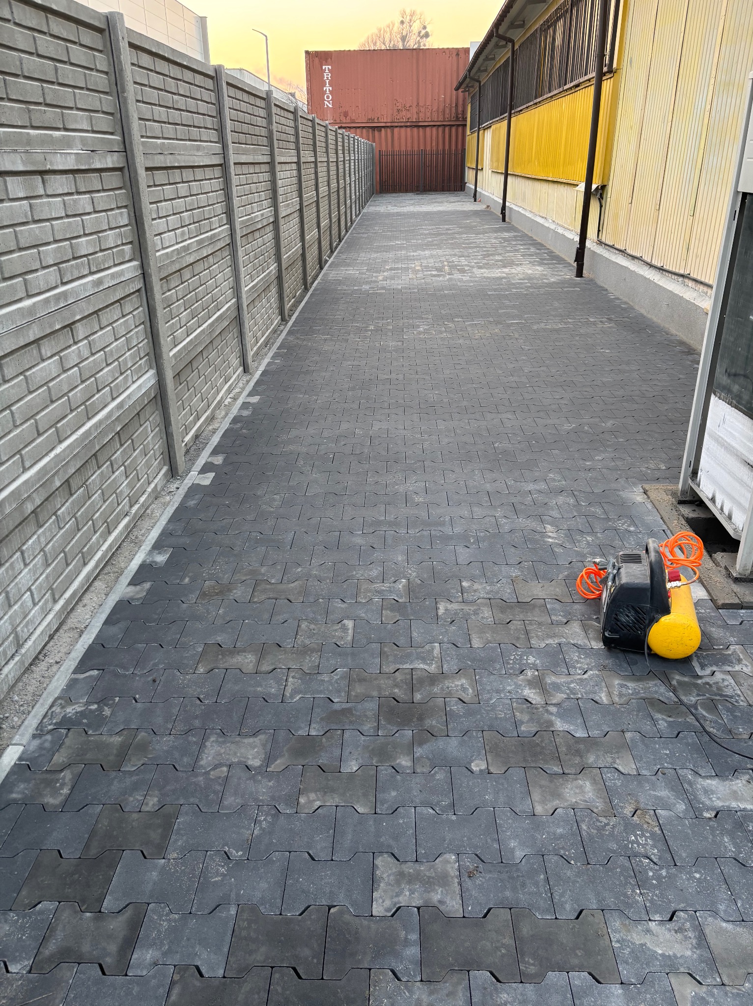 Ułożona szara kostka brukowa typu 'H' na posesji, widoczny fragment betonowego ogrodzenia i żółty kompresor z pomarańczowym przewodem.