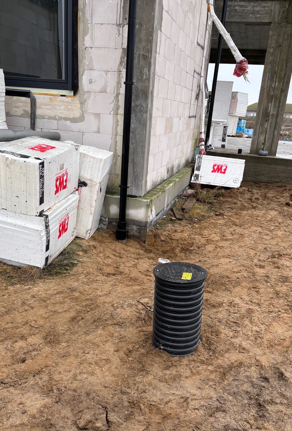 Czarna studzienka kanalizacyjna z żółtą naklejką na piasku obok fundamentów domu z białego bloczka, izolacji termicznej i rury spustowej.