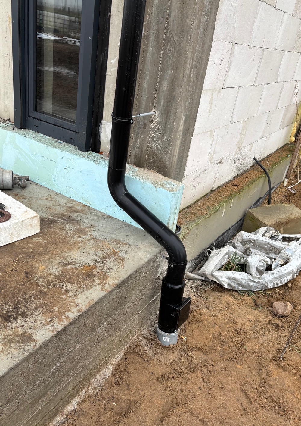 Czarna rura spustowa zamontowana przy fundamencie budynku z bloczków, widoczna izolacja fundamentu, ziemia i fragment okna.