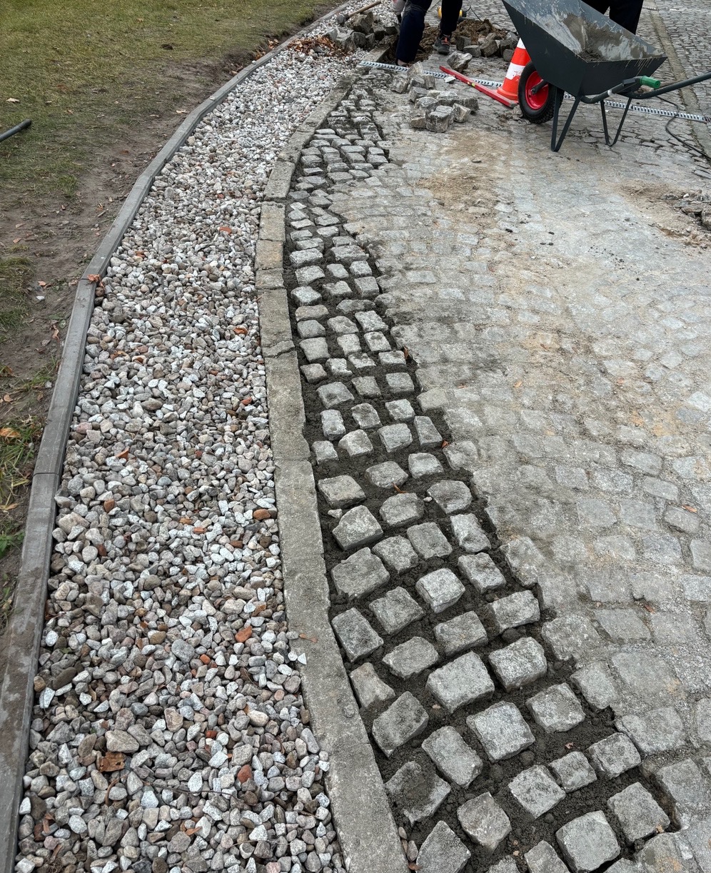 Układanie kostki brukowej: fragment chodnika z szarej kostki, częściowo ułożonej, z widoczną warstwą podsypki, obok obsypka z drobnych kamieni, w tle taczka i robotnicy.