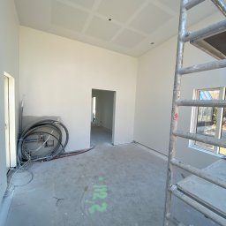 Mateusz Antczak - Wnętrze w trakcie prac wykończeniowych: białe ściany, podwieszany sufit z perforacją, instalacje, drabina malarska. Widok na sąsiednie pomieszczenie.