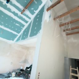 Mateusz Antczak - Wnętrze w trakcie remontu: sufit z płyt gipsowo-kartonowych na drewnianej konstrukcji, częściowo otynkowane ściany, drabina i narzędzia w tle. Prace wykończeniowe.