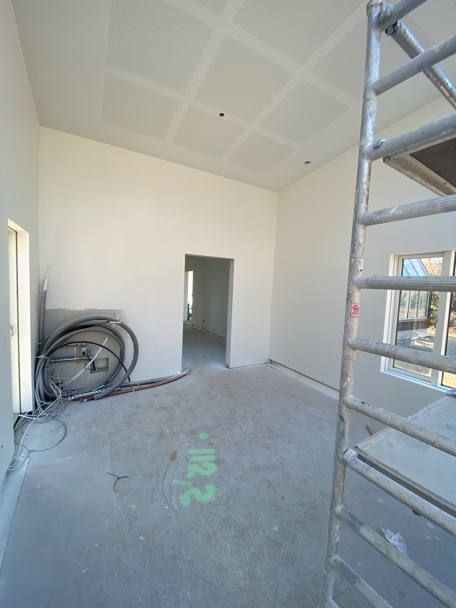 Wnętrze w trakcie prac wykończeniowych: białe ściany, podwieszany sufit z perforacją, instalacje, drabina malarska. Widok na sąsiednie pomieszczenie.