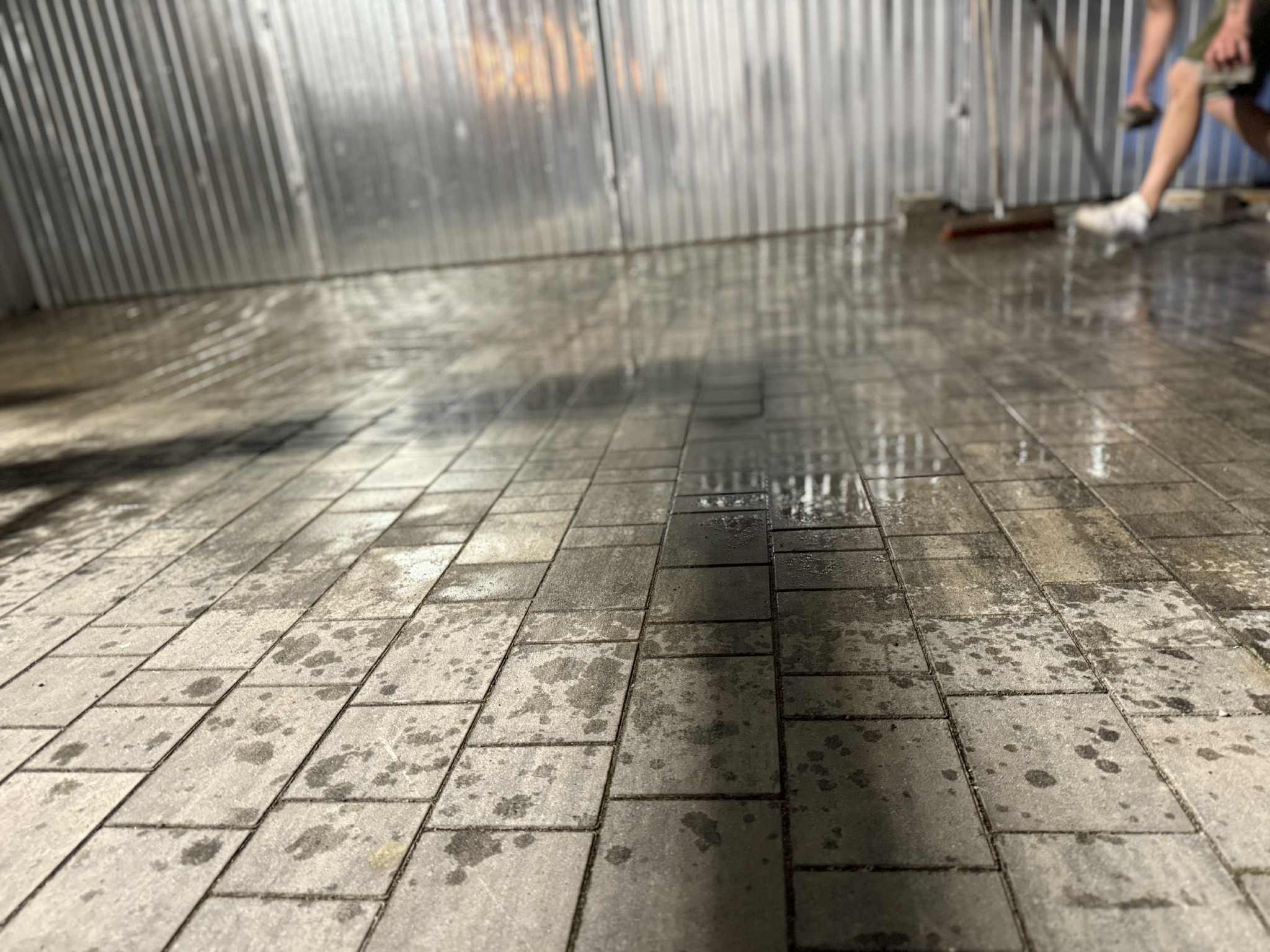 Mycie kostki brukowej w garażu: mokra powierzchnia z ciemnymi plamami, osoba w tle z miotłą. Metalowa, pofalowana ściana w tle odbija światło.