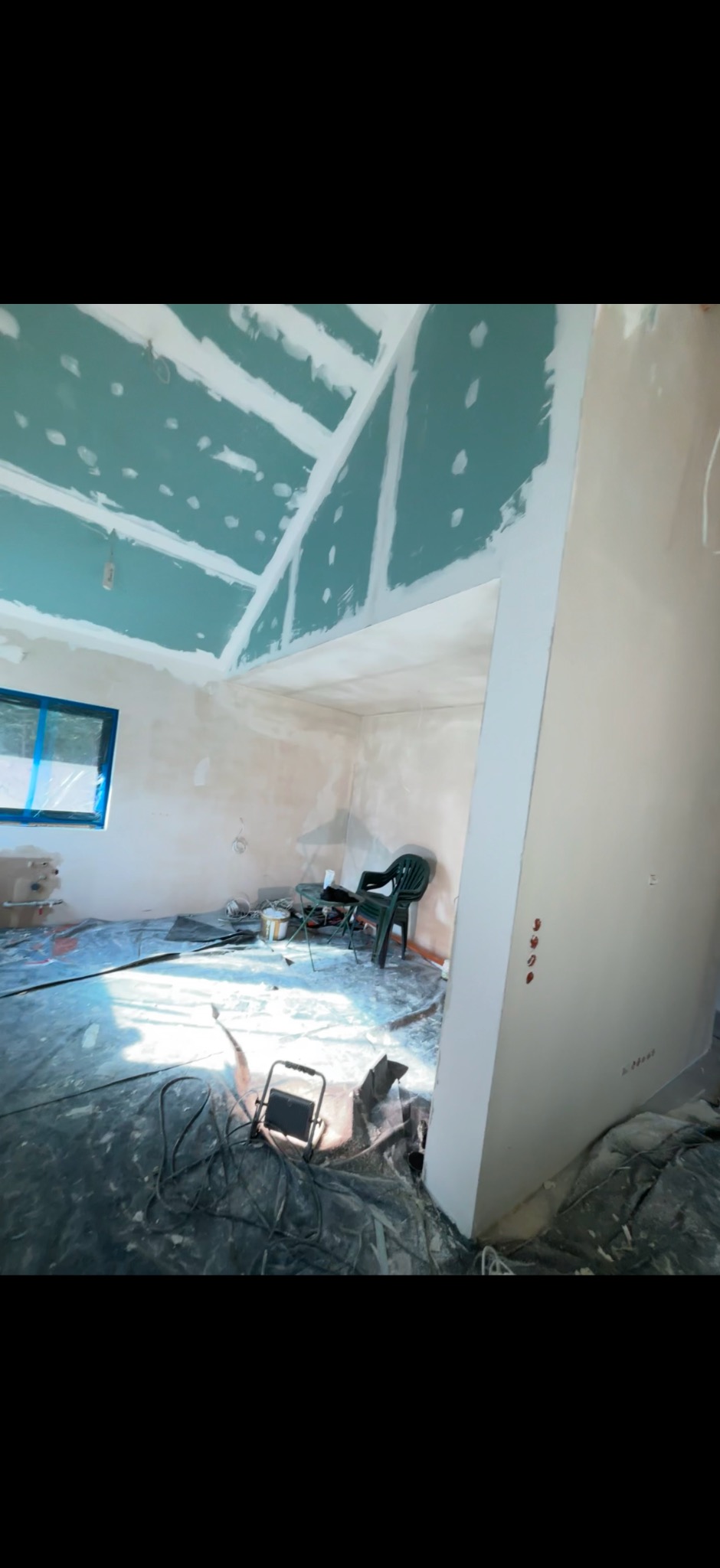 Wnętrze w trakcie remontu: sufit z płyt gipsowo-kartonowych po szpachlowaniu, zabezpieczona folią podłoga, narzędzia i lampa budowlana. Widok na pokój przez otwarty otwór.