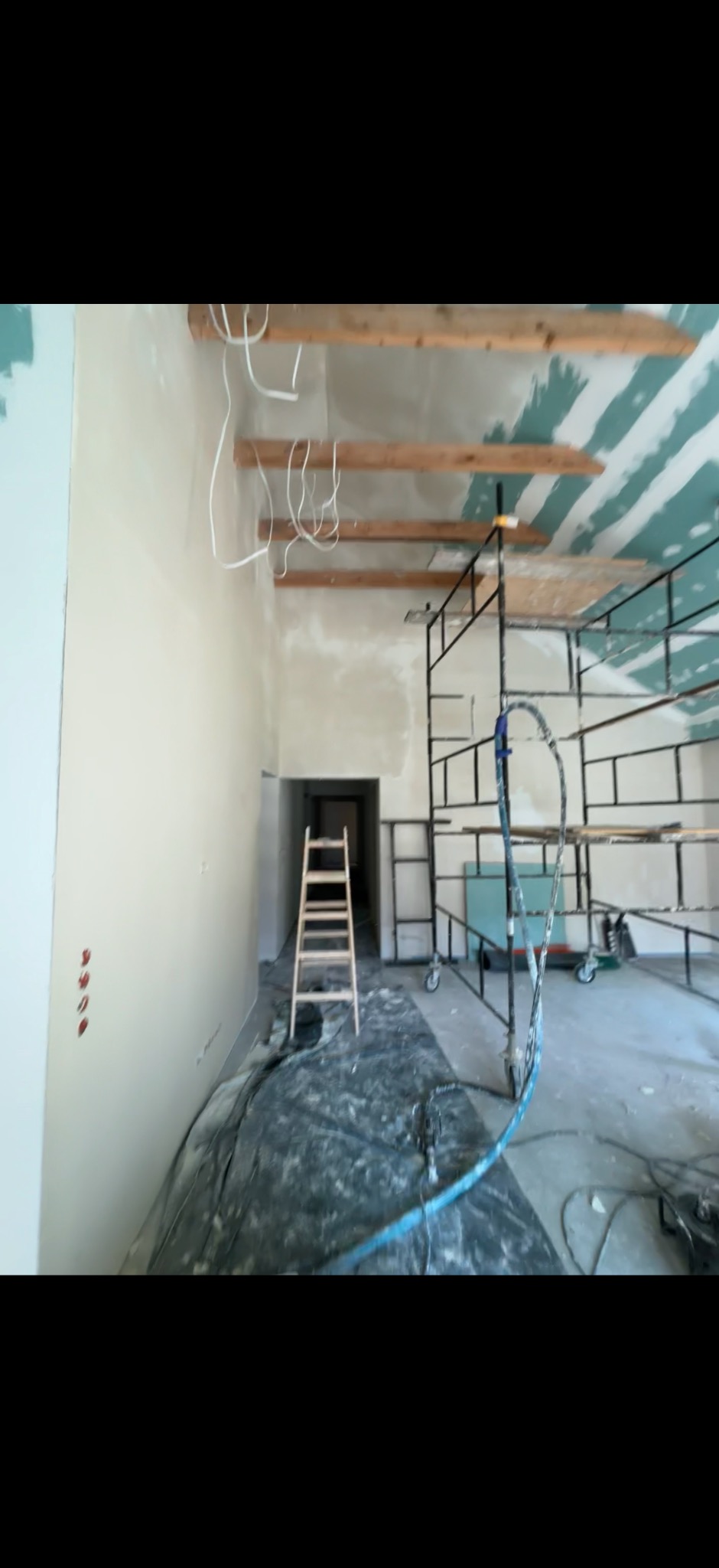 Wnętrze w trakcie remontu: ściany po szpachlowaniu, widoczne przewody elektryczne pod sufitem, rusztowanie i drabina. Przygotowanie do dalszych prac wykończeniowych.