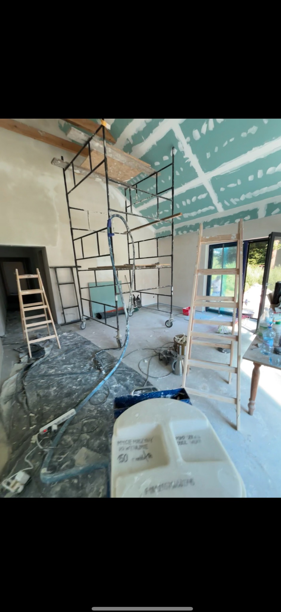 Wnętrze w trakcie szpachlowania: widoczne rusztowanie, drabina, narzędzia i częściowo wykończone ściany z płyt gipsowo-kartonowych. Detal: pojemnik z napisem 'Mycie maszyn'