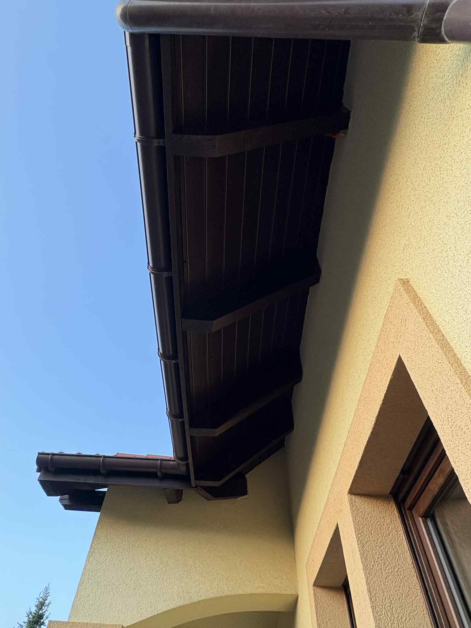 Elewacja budynku z brązowymi rynnami i podbitką, widok z dołu na tle błękitnego nieba. Teksturowana, beżowa ściana z narożnikiem okna. Detal architektoniczny.