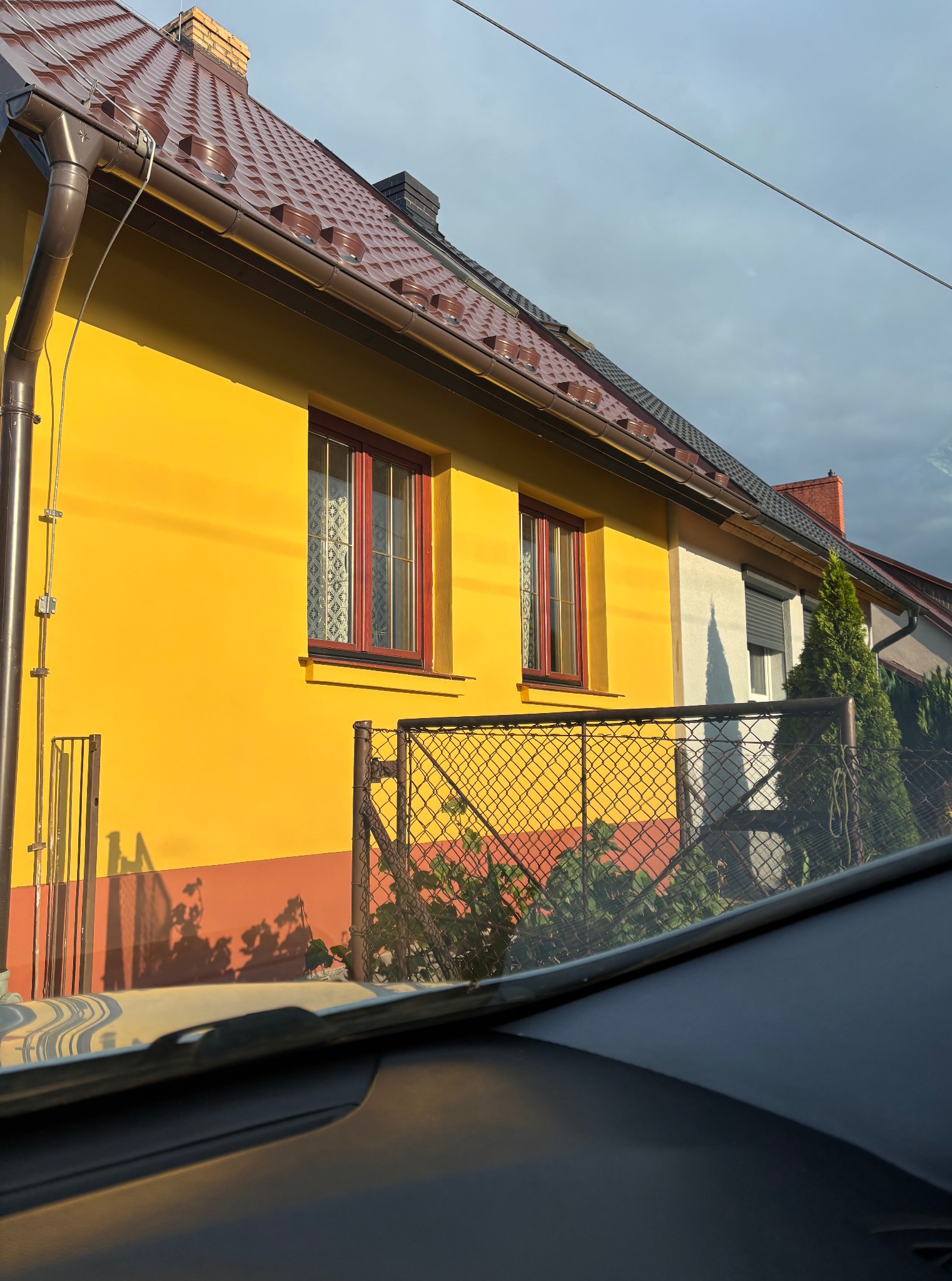 Żółty dom z brązowym dachem i czerwonymi ramami okiennymi, widziany zza szyby samochodu, cień roślin na ścianie.