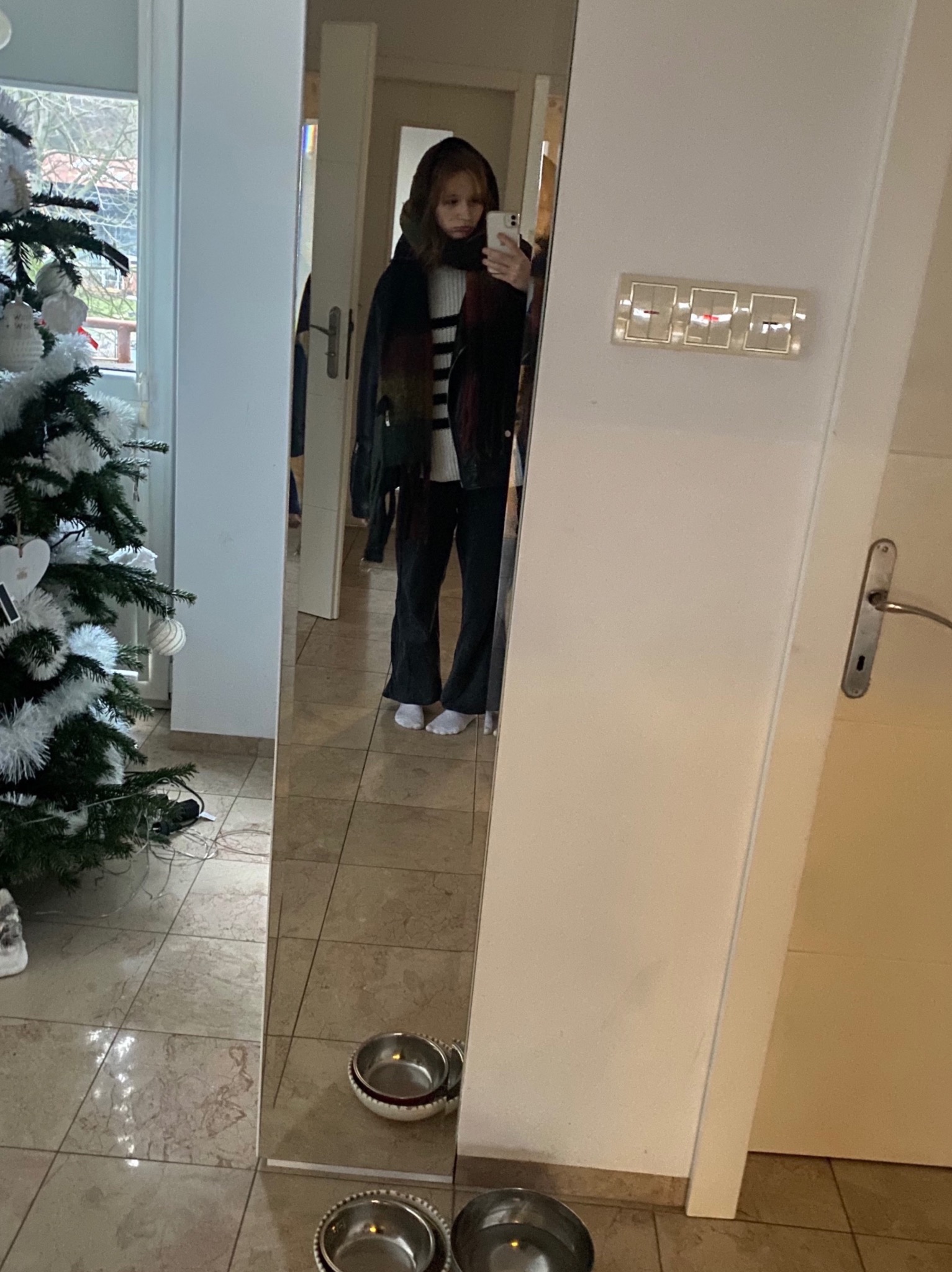 Odbicie w lustrze przedstawia dziewczynę robiącą sobie zdjęcie telefonem w przedpokoju z choinką, widoczne miski dla zwierząt na podłodze.